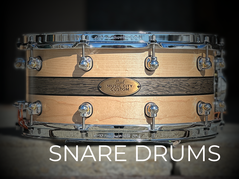 A close-up of a snare drum with a natural wood finish and metal hardware, featuring a plaque labeled "Music City Custom." Text at the bottom reads "SNARE DRUMS."